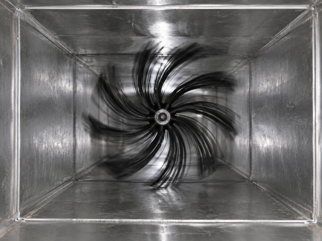 View of an industrial ventilation duct with a spinning fan inside, showcasing the metal interior and motion blur of the fan blades.